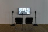 Installation view of a video work. A television monitor is attached to the wall, flanked by two speakers on narrow stands. In front of the monitor are two black chairs with silver metal legs.
