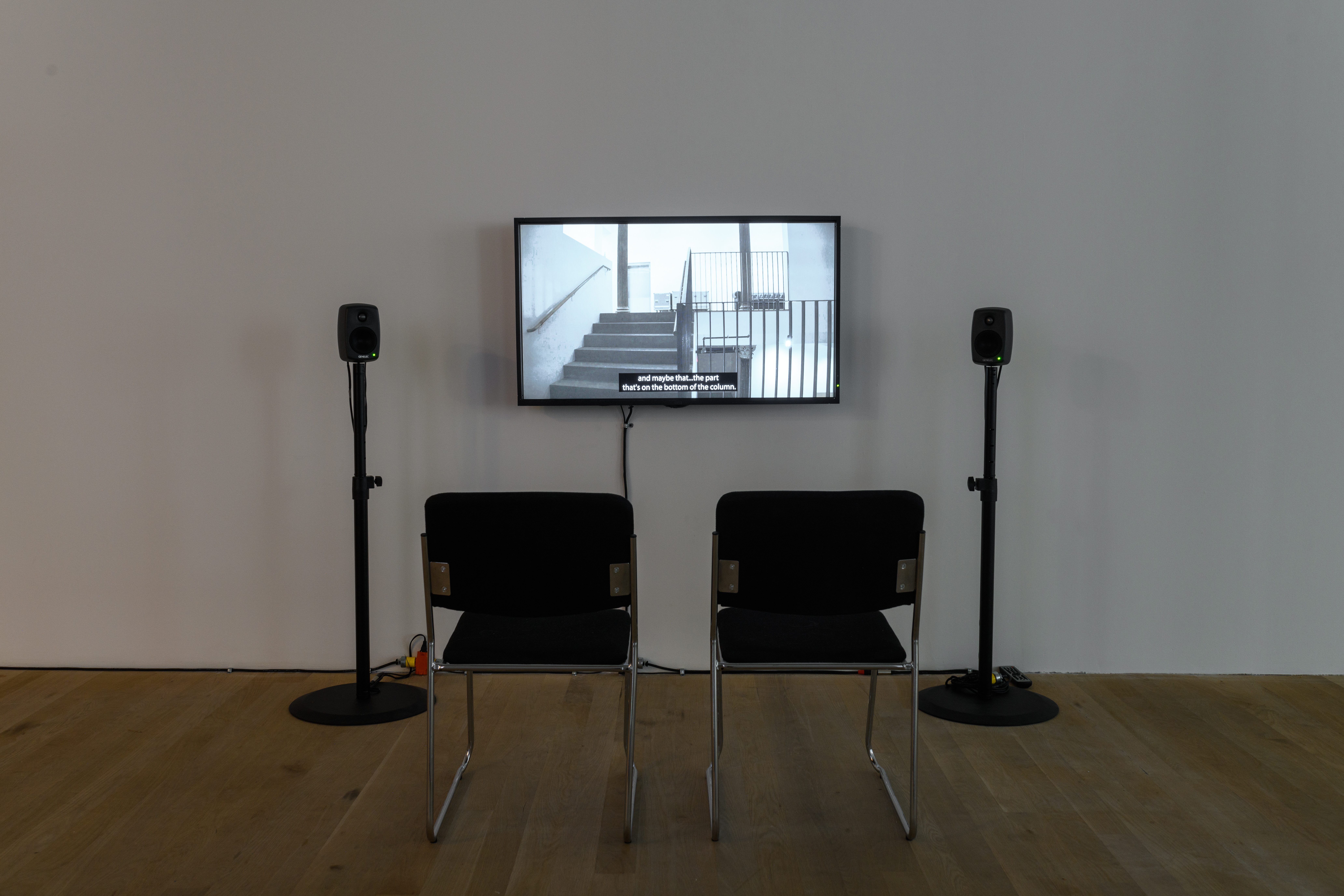 Installation view of a video work. A television monitor is attached to the wall, flanked by two speakers on narrow stands. In front of the monitor are two black chairs with silver metal legs.
