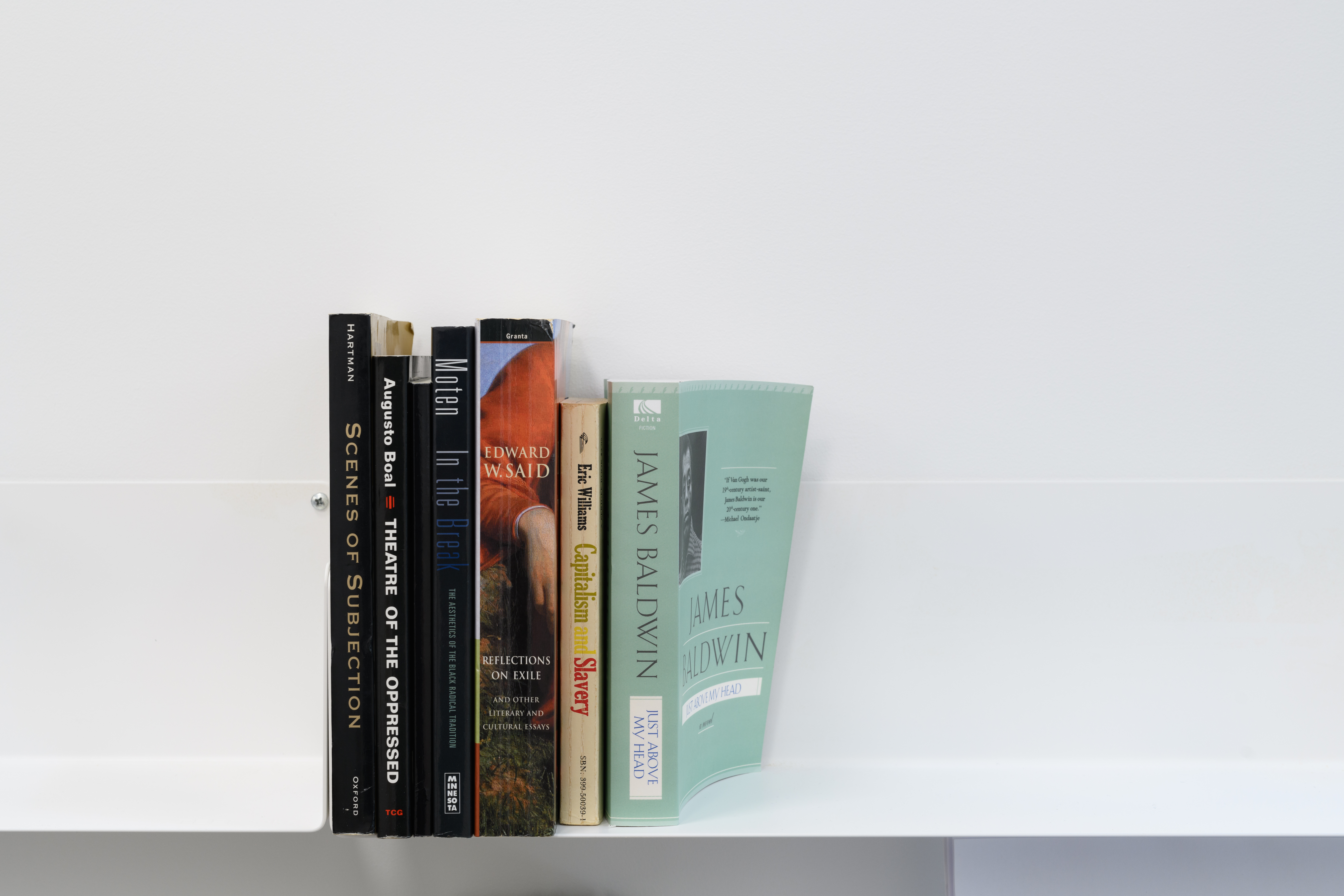 Six books on a white shelf against a white wall. From left, their authors are Saidiya Hartman, Augusto Boal, Fred Moten, Edward W. Said, Eric Williams, and James Baldwin.