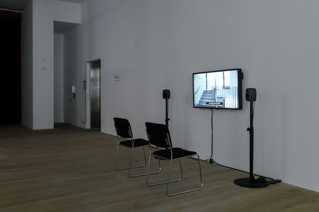 Installation view of a video work. A television monitor is attached to the wall, flanked by two speakers on narrow stands. In front of the monitor are two black chairs with silver metal legs. In the background, a dark doorframe and elevator are visible.