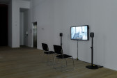 Installation view of a video work. A television monitor is attached to the wall, flanked by two speakers on narrow stands. In front of the monitor are two black chairs with silver metal legs. In the background, a dark doorframe and elevator are visible.