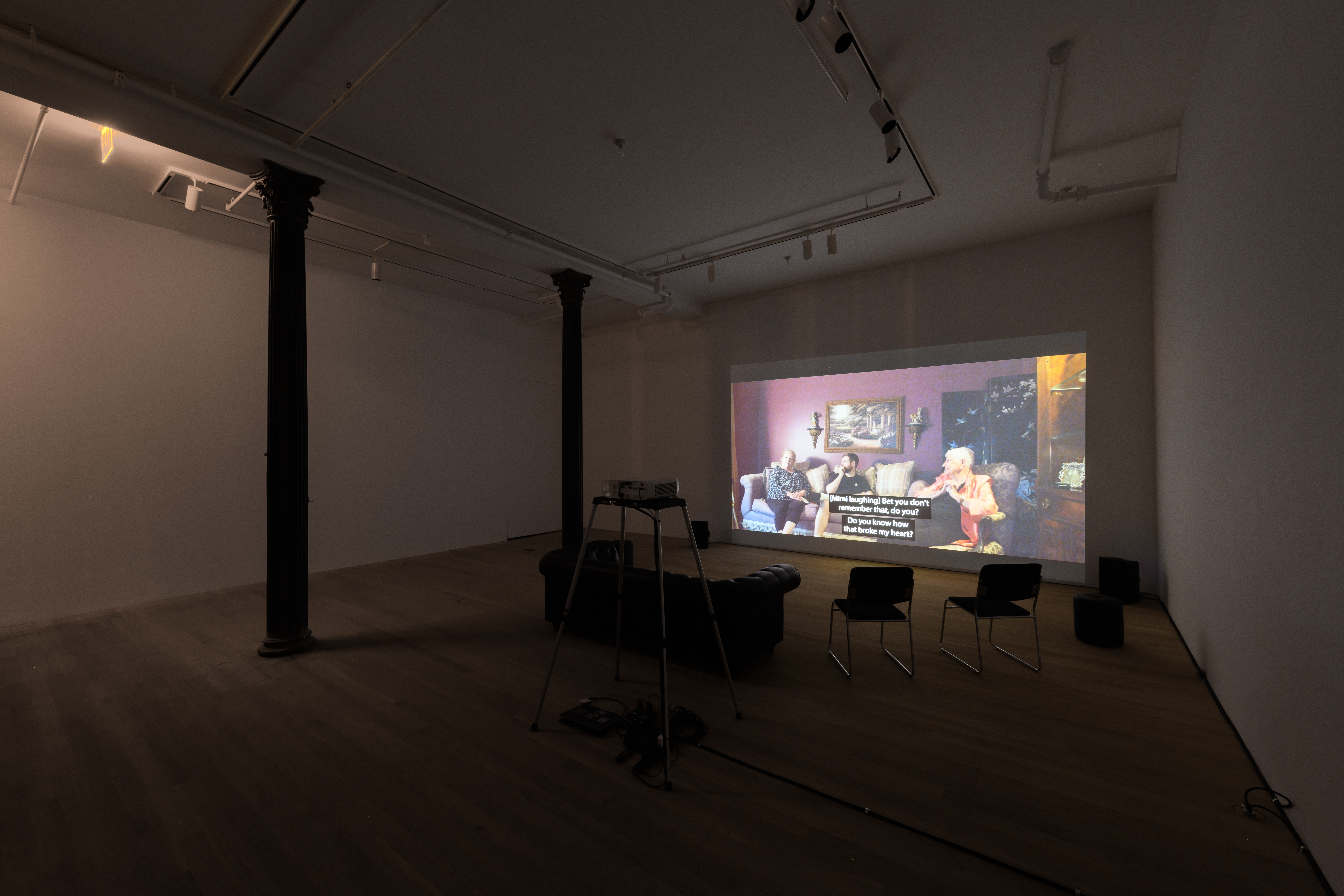 A darkened room with a projected video, several chairs and a black sofa. Two colums are visible on the left.