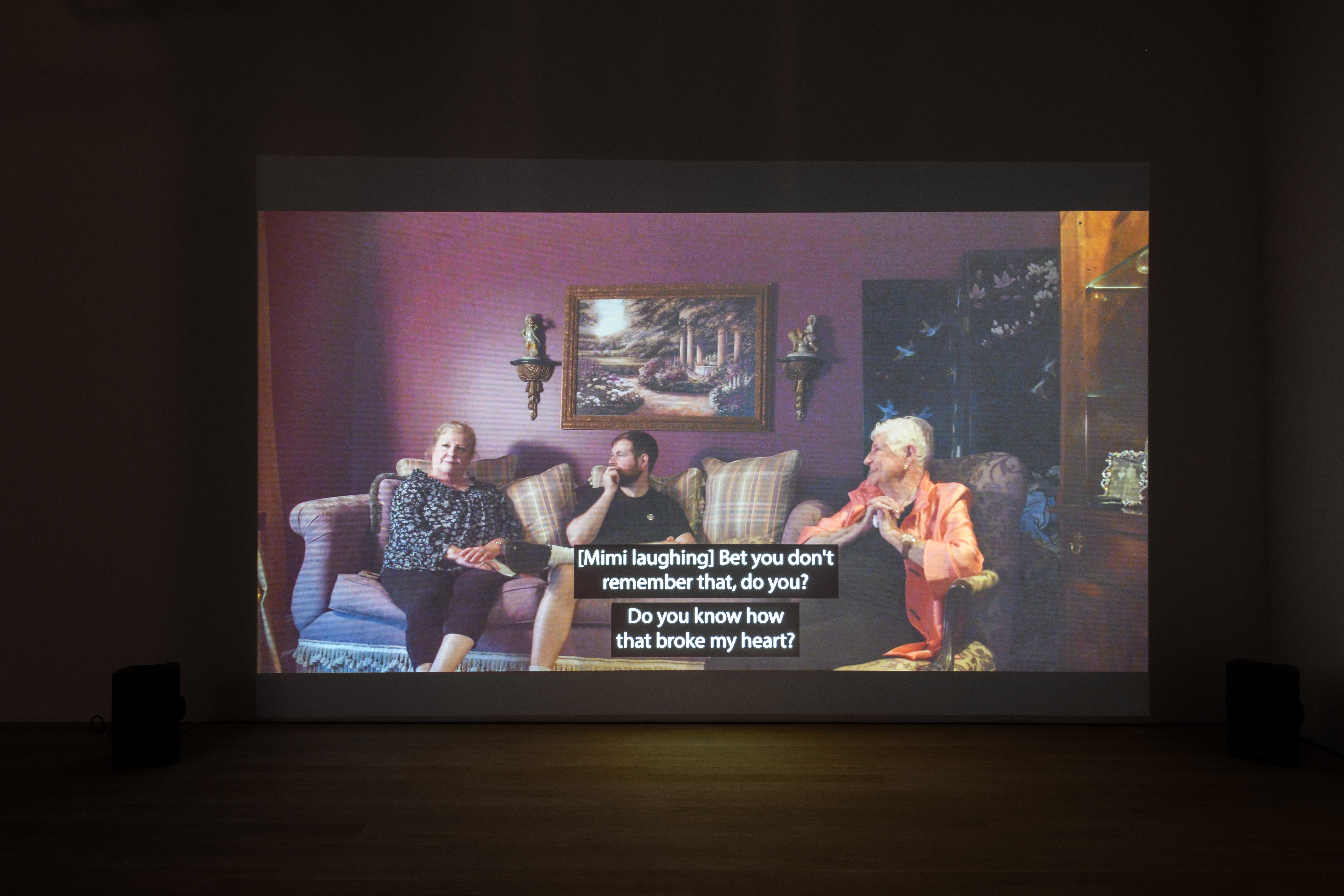 Head on view of a projected video. The screen shows three figures sitting and includes closed captioning that reads 