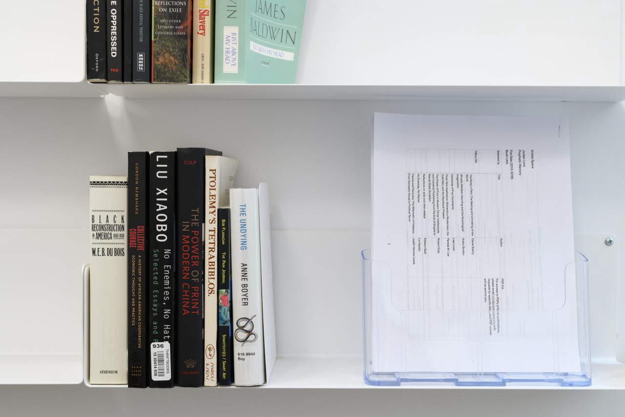 A set of white metal shelves against a white wall. The top shelf hold seven books and the bottom shelf holds seven books and stapled pamphlets in a clear stand.