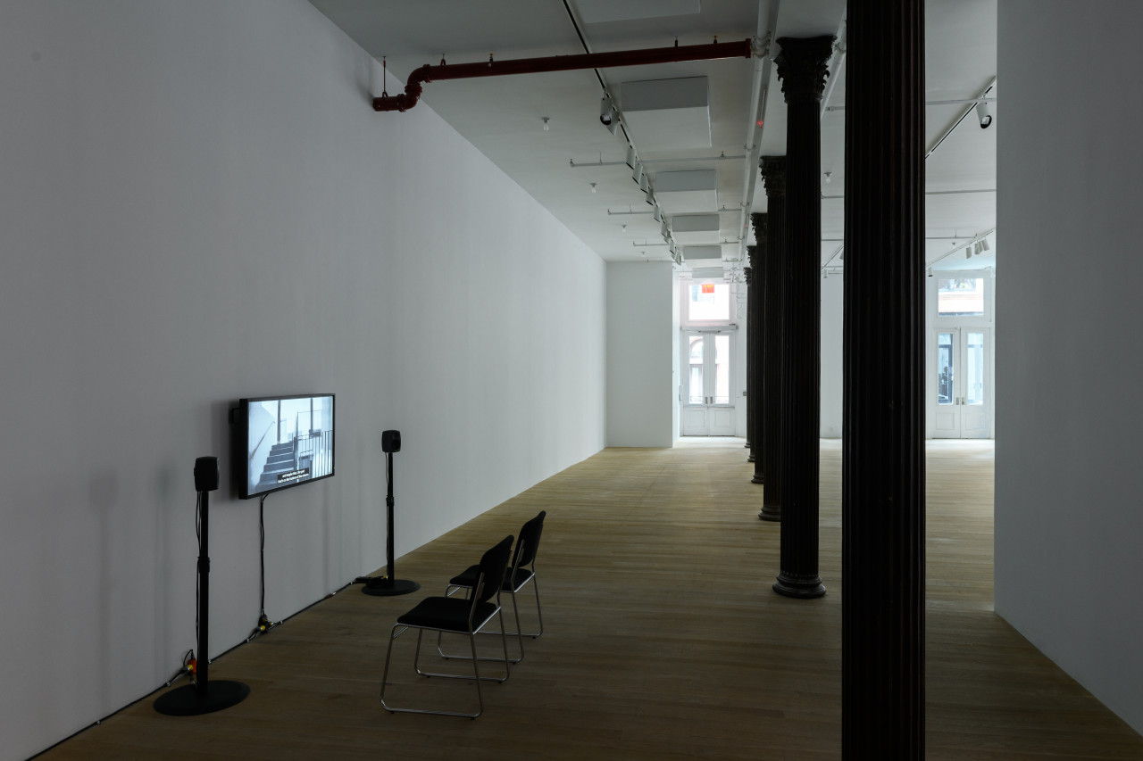 Installation view of a video work. A television monitor is attached to the wall, flanked by two speakers on narrow stands. In front of the monitor are two black chairs with silver metal legs. On the right, a row of columns extends into the background towards sunlit windows.