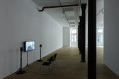 Installation view of a video work. A television monitor is attached to the wall, flanked by two speakers on narrow stands. In front of the monitor are two black chairs with silver metal legs. On the right, a row of columns extends into the background towards sunlit windows.
