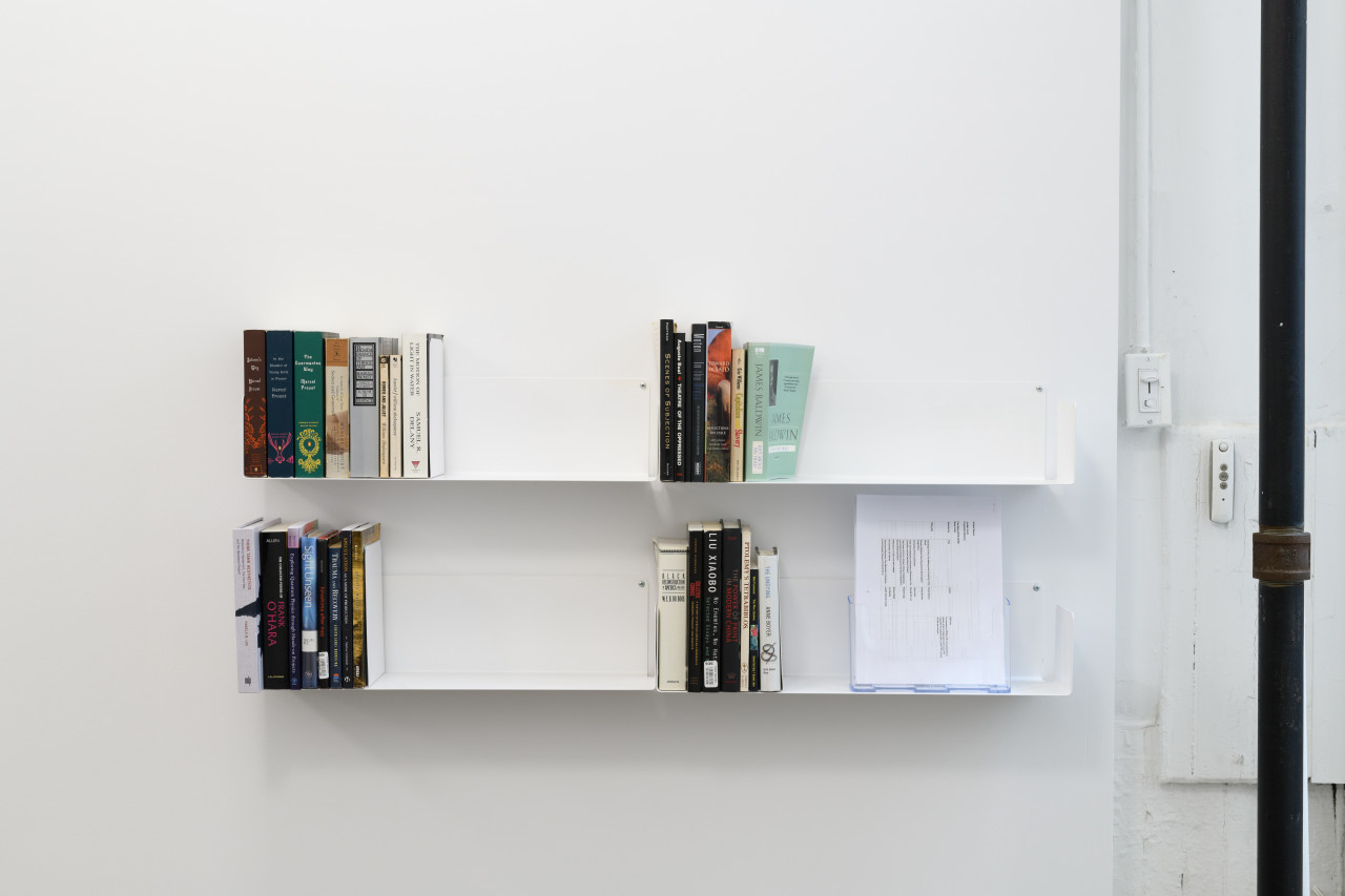 Two white shelves attached to a white wall hold four sets of books and some papers. On the right is a black metal pole.