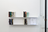 Two white shelves attached to a white wall hold four sets of books and some papers. On the right is a black metal pole.