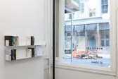A view of a gallery space with white walls and a window looking out onto a street. On the left wall two shelves hold various books and papers.