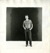 A figure standing in front of a black square canvas in a gallery space.