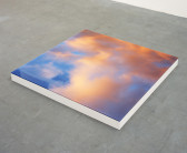 A low platform on a concrete floor supporting a glasstop photograph of pink clouds on a blue sky.