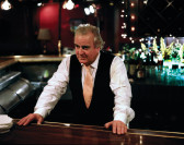 An older man in a white collared shirt, pink tie, and black vest leans against a shining countertop—perhaps a bar—with both hands. A restaurant interior and fishtank gleam behind him.