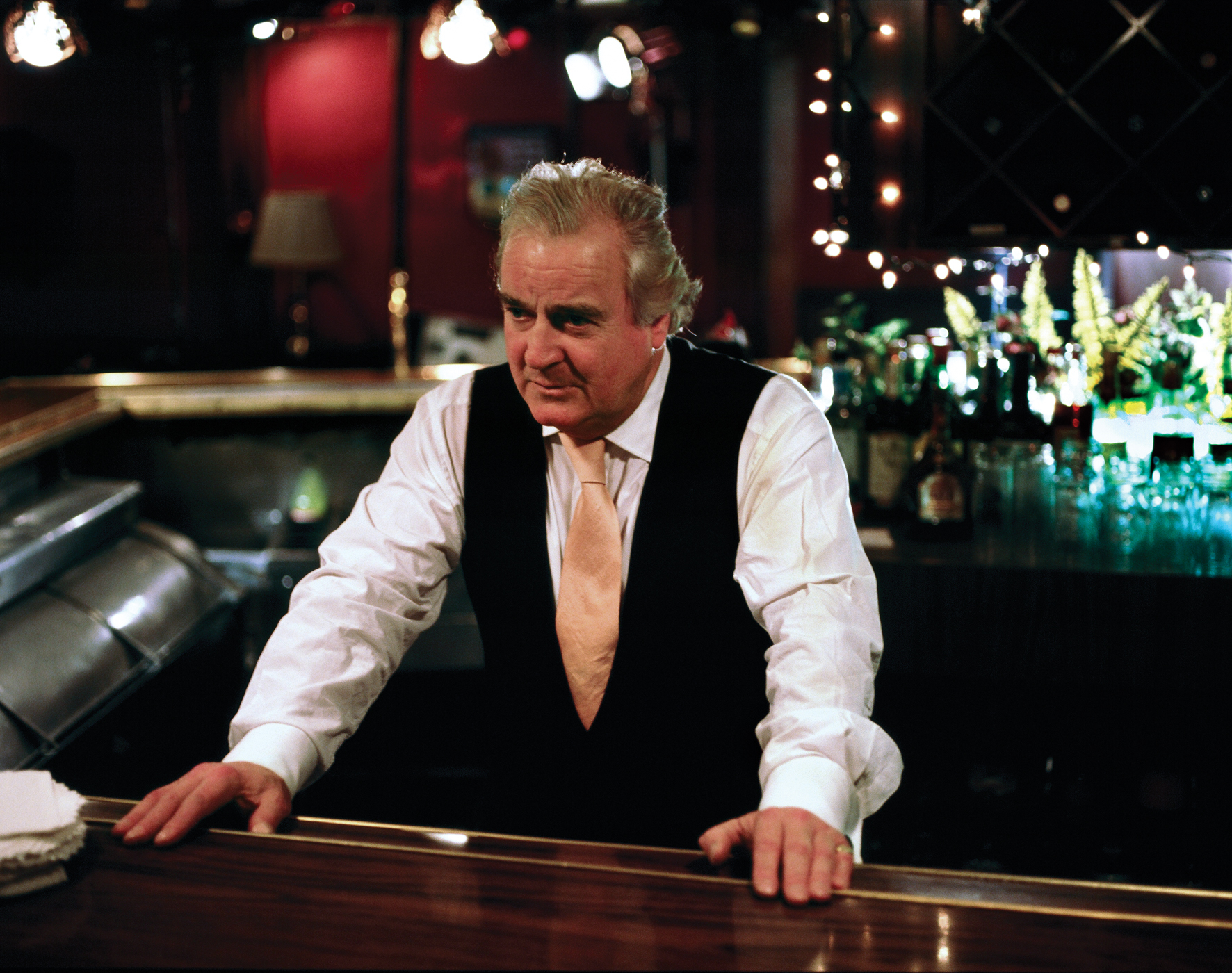 An older man in a white collared shirt, pink tie, and black vest leans against a shining countertop—perhaps a bar—with both hands. A restaurant interior and fishtank gleam behind him.