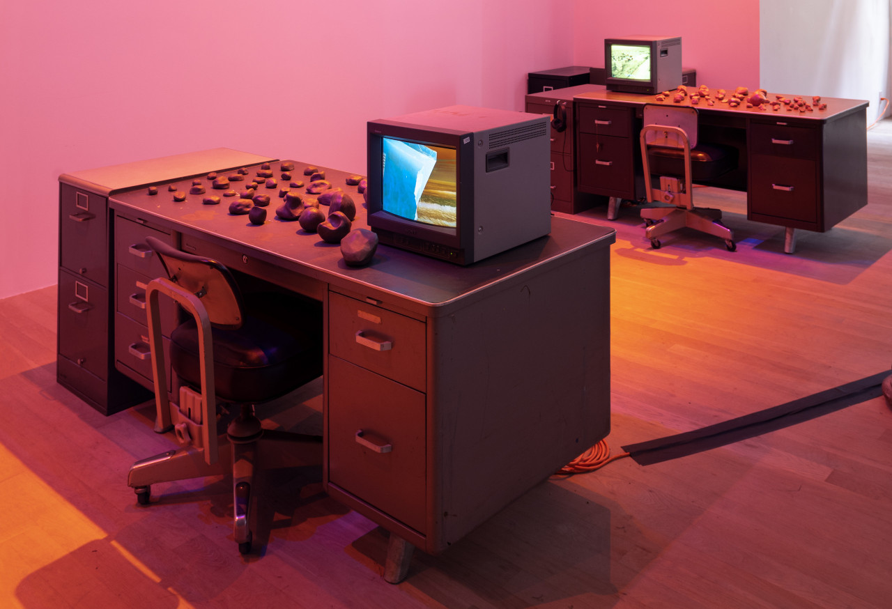 Image of two desks, each with a chair, TV monitor playing a video, and clay rocks placed on top.