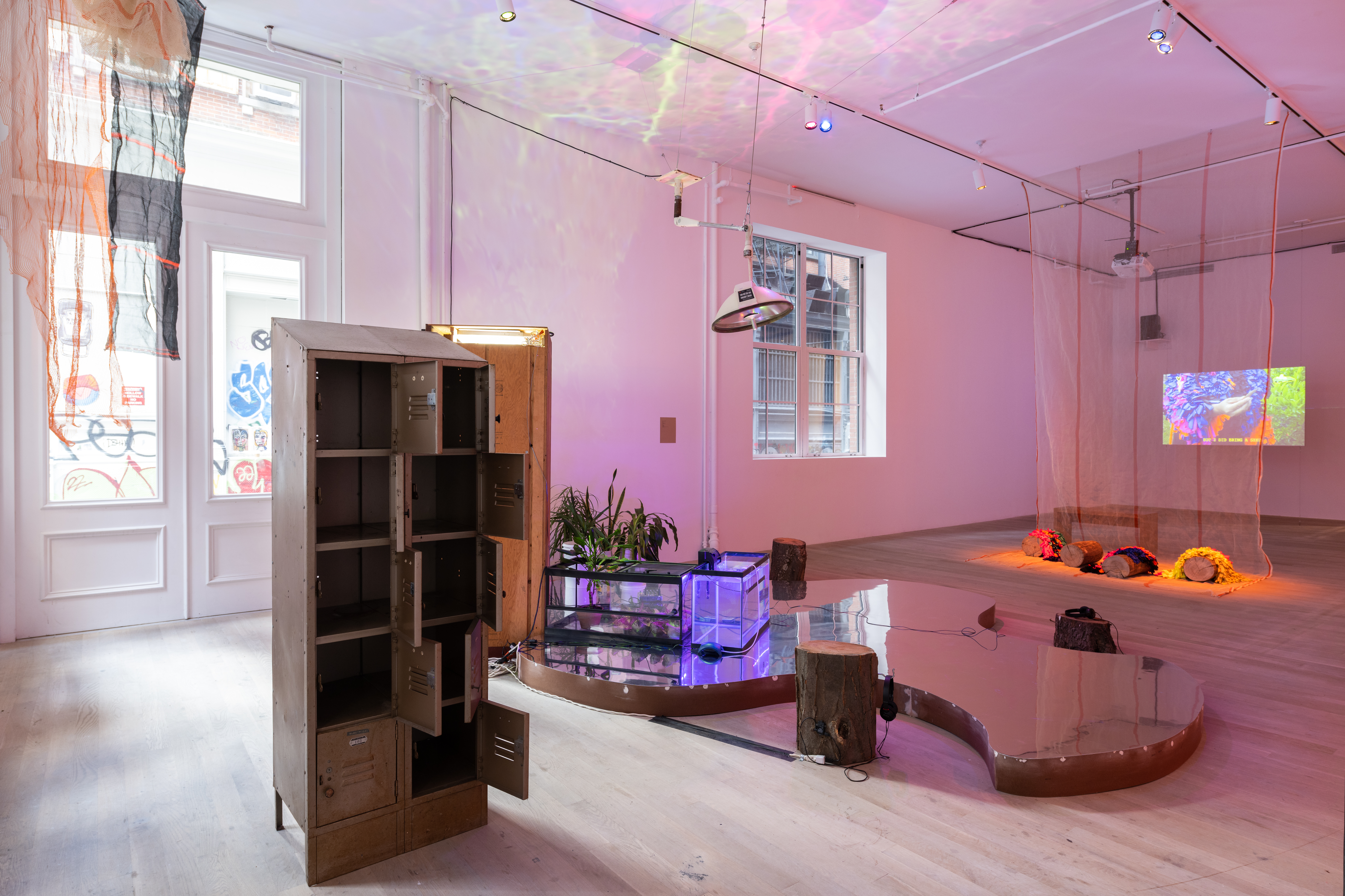 Image of a gallery space with a metal locker, mirrored wooden base, and some tree stumps and plants. A large light and construction nets hang from the ceiling, and on the wall in the back a video is projected onto a screen.