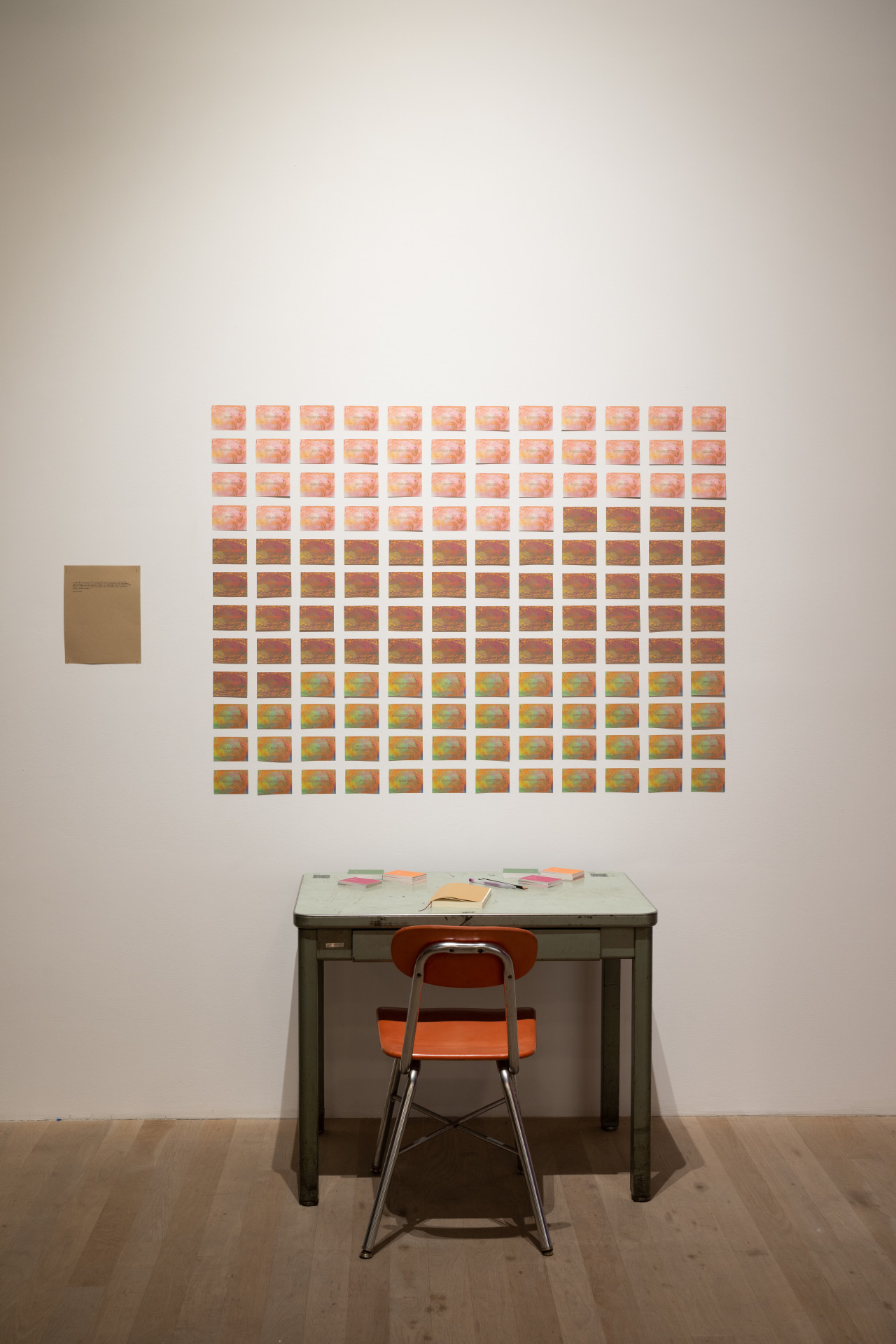 A chair and desk face a white wall which has a grid of cards in varying colors on it. A notebook, stacks of cards, and pens are on the desk.