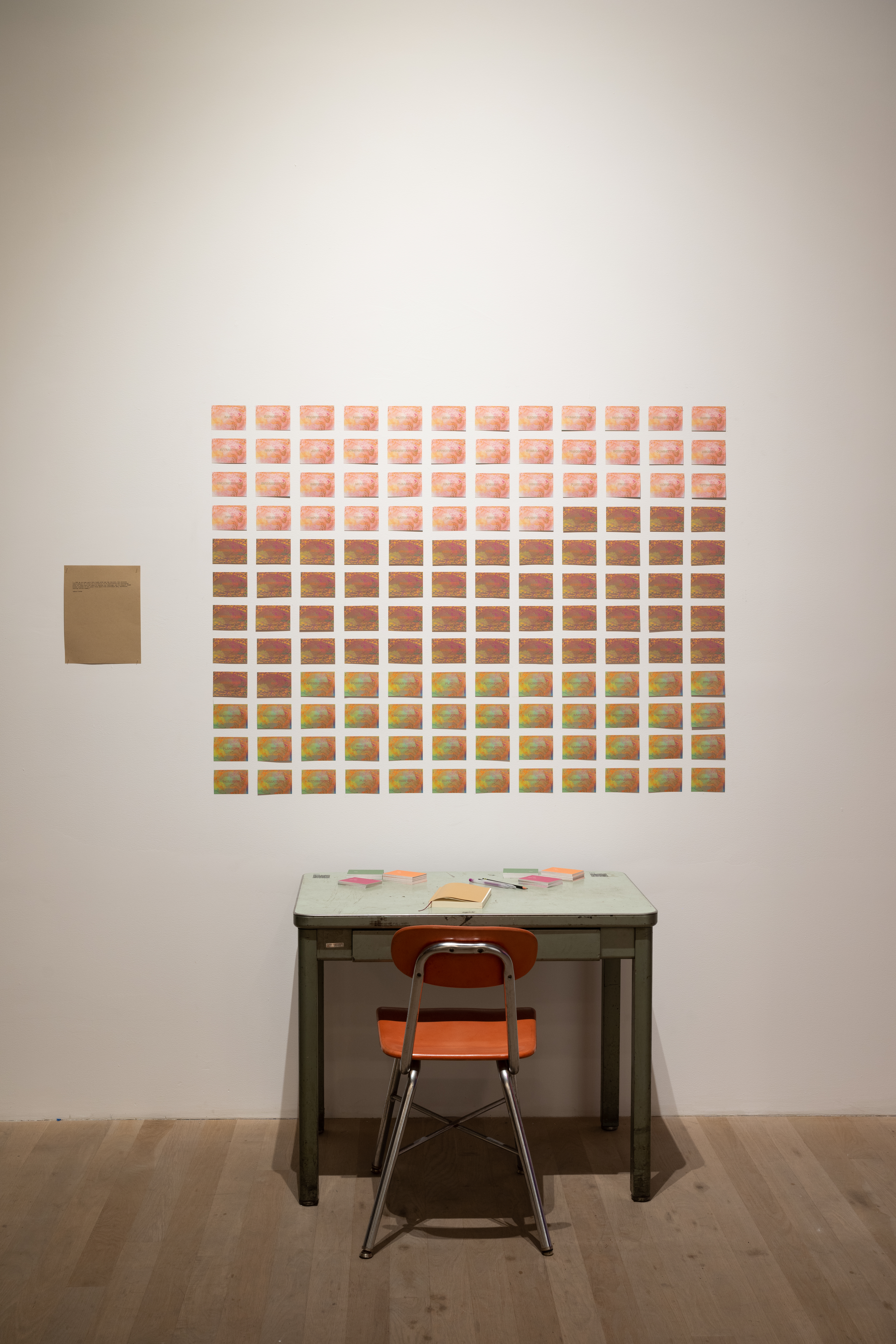 A chair and desk face a white wall which has a grid of cards in varying colors on it. A notebook, stacks of cards, and pens are on the desk.