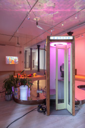 A picture of a gallery space with a phonebooth in the foreground. Behind the phonebooth is a reflective wooden base, and some plants. There is a video projected on a wall in the back of the space.