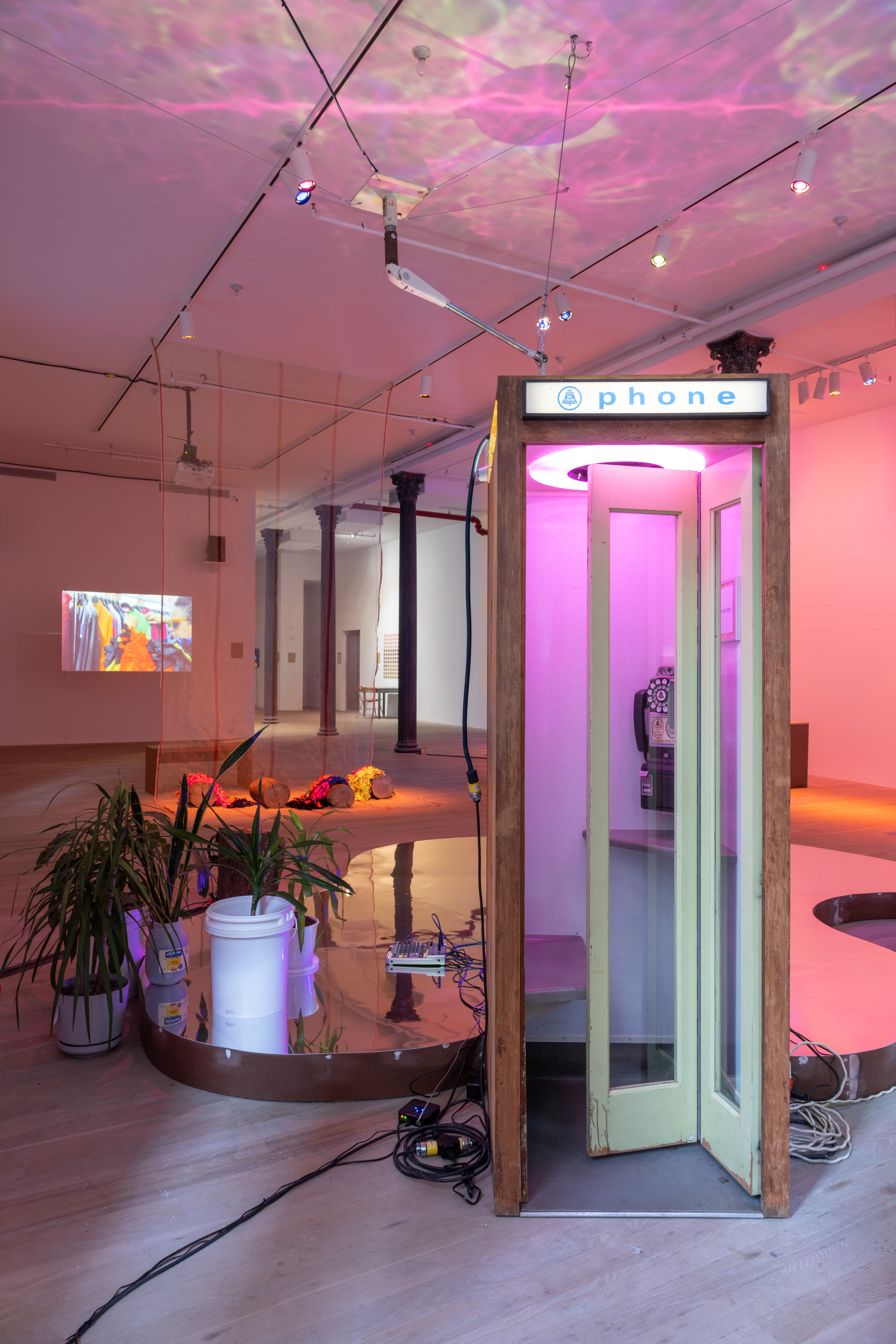 A picture of a gallery space with a phonebooth in the foreground. Behind the phonebooth is a reflective wooden base, and some plants. There is a video projected on a wall in the back of the space.