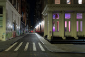 Image of the street outside the gallery, looking into an alleyway with a white pillared building on the right with purple light showing behind the windows.