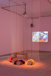 A construction net hangs from the ceiling to the floor with four tree trunks placed on top of it on the floor. Colorful fabrics are placed on top of the tree trunks. A wooden bench sits behind the trunks and faces a video projection in front of the wall.