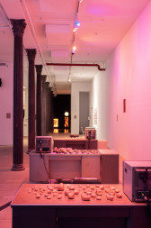 Three desks are lined up in front of each other, each desktop has a TV monitor and clay rocks. In the back is a dark room with yellow LED light sculptures.