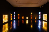A large gallery room's walls are painted black, with a black vinyl floor, and white ceiling. Along the walls are yellow and orange LED lighting sculptures that have dried vegetable leather stitched on top of the lights. In the corners of the room three TVs playing animations of rushing water are placed on top of each other.