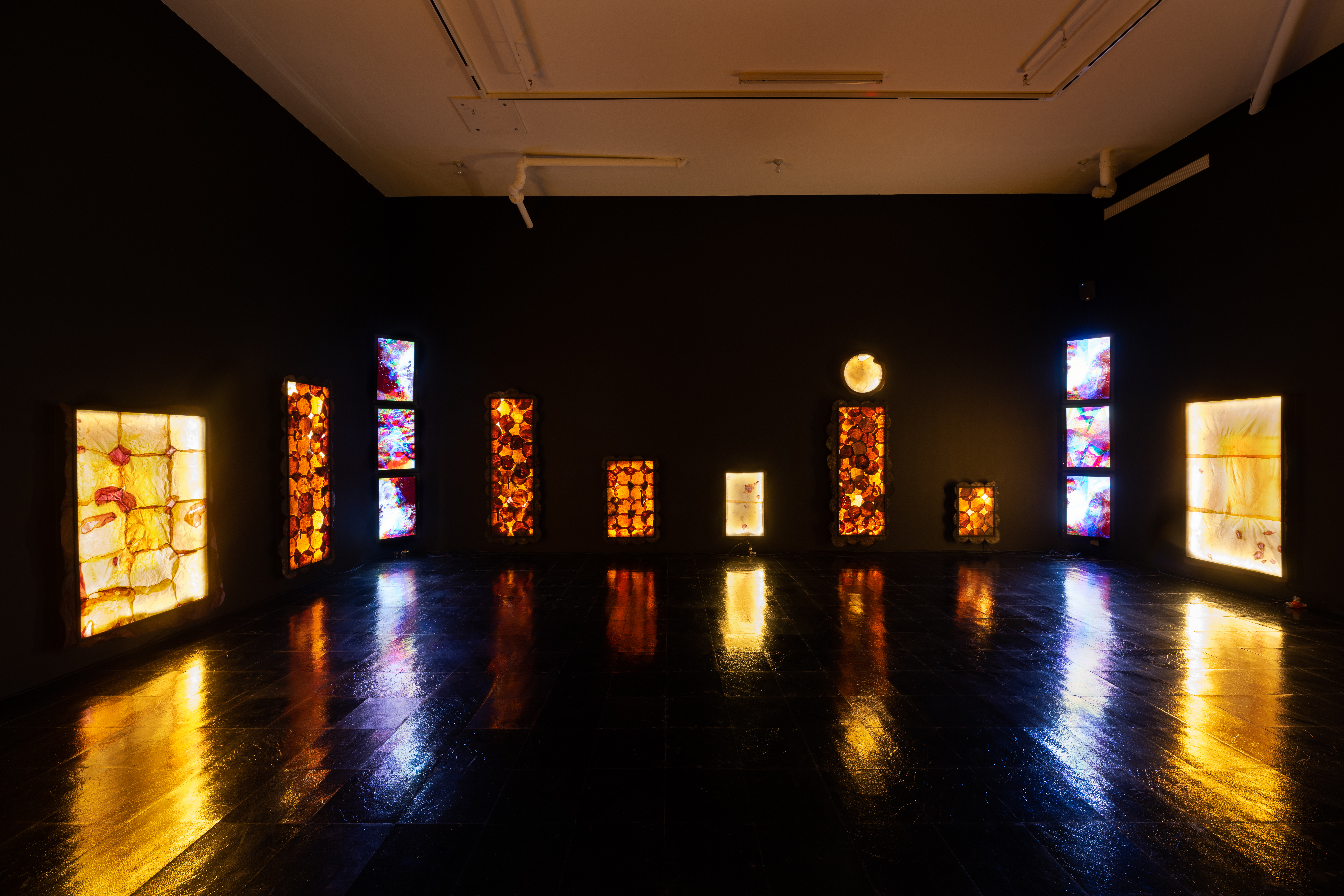 A large gallery room's walls are painted black, with a black vinyl floor, and white ceiling. Along the walls are yellow and orange LED lighting sculptures that have dried vegetable leather stitched on top of the lights. In the corners of the room three TVs playing animations of rushing water are placed on top of each other.