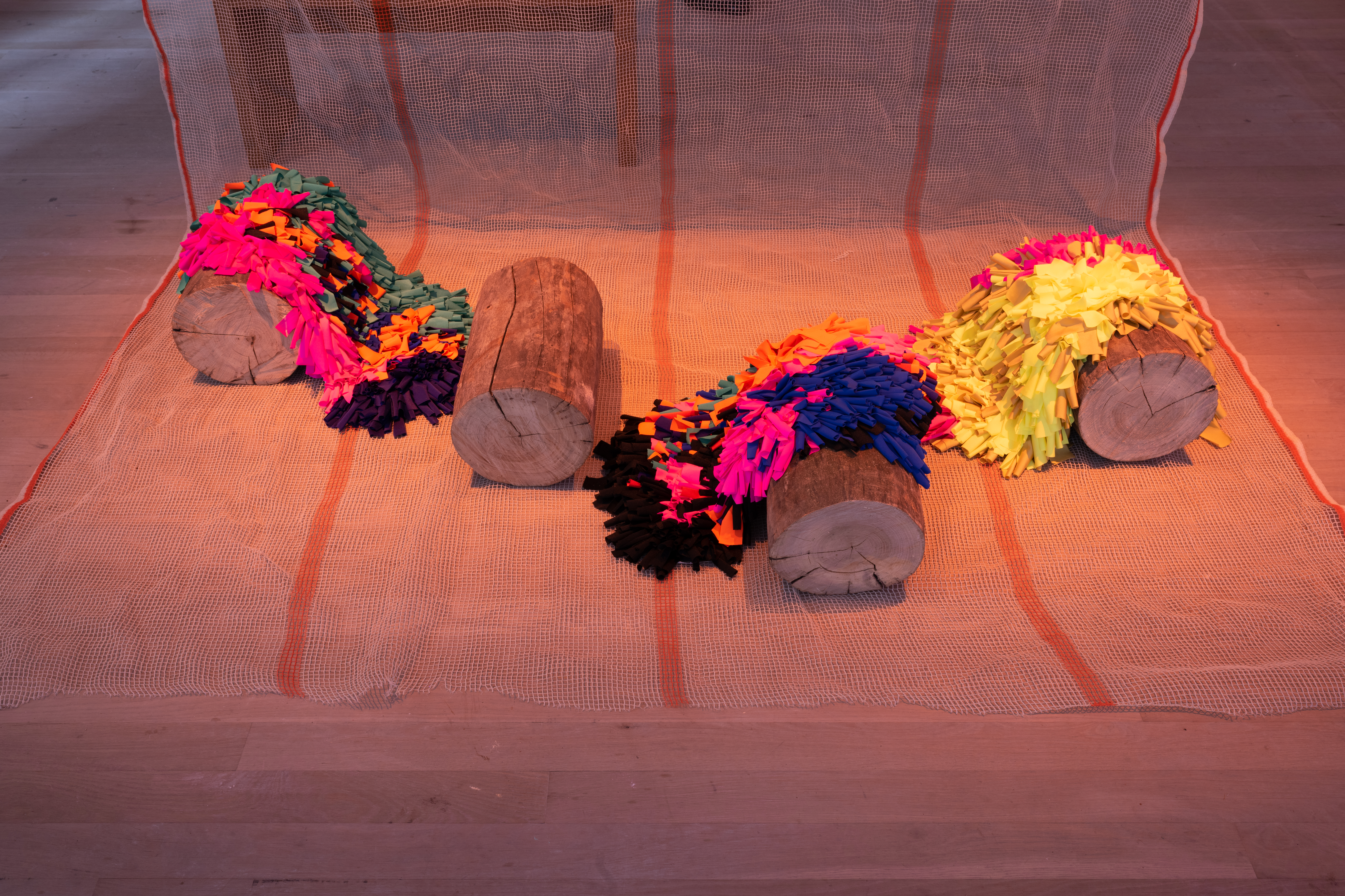 Inside an orange-hued room are four tree trunks sitting atop a striped construction net. Three trunks are adorned in purple, orange, blue, pink, yellow, and green fabrics. A fourth trunk sits without fabric.