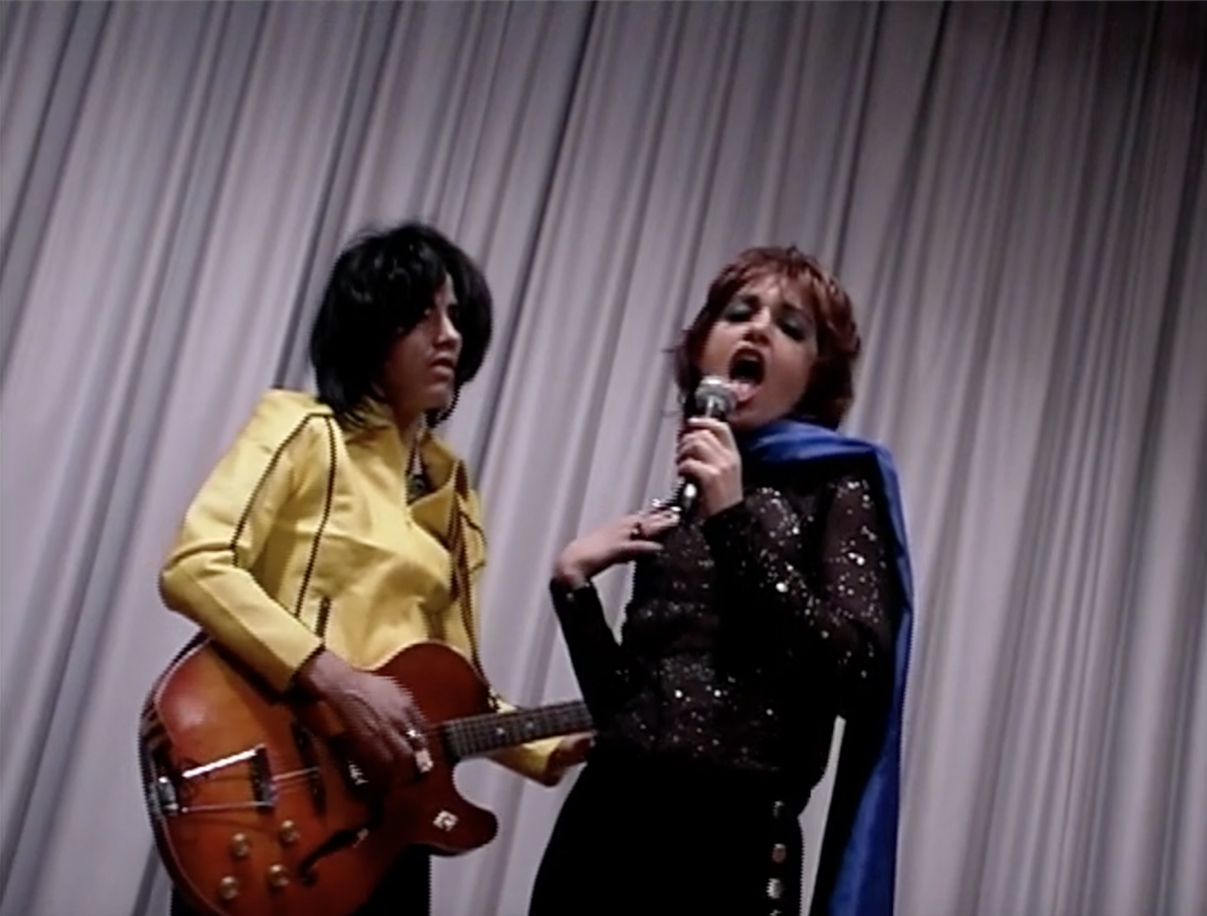 An upward angled view of two people performing with a gray curtain behind them. One person, with black hair and a yellow jacket, is playing an electric guitar, the other, with red hair and wearing a black sequin top and blue scarf with matching eyeshadow, is singing into a mic.