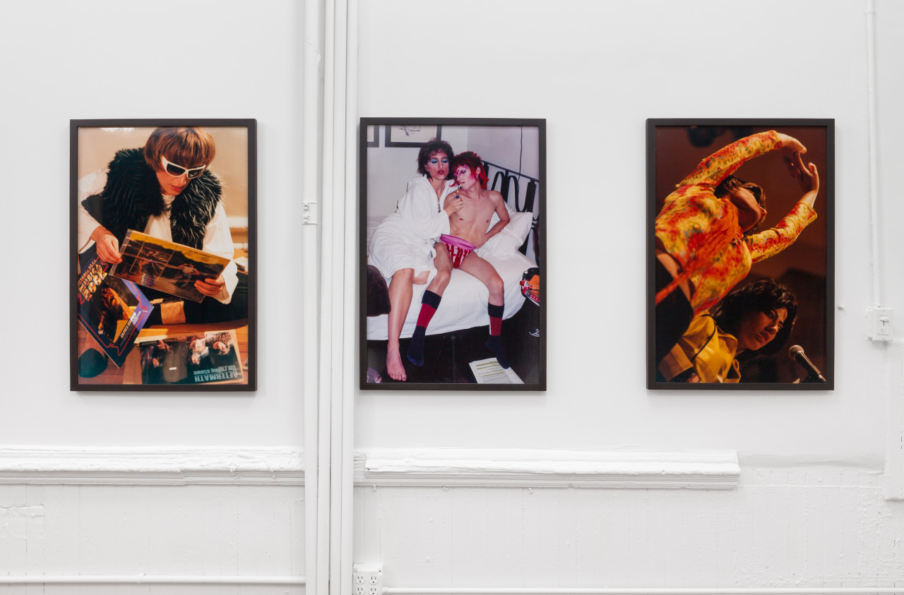 Three vertical prints, framed and lined on a white wall. From left to right: A person wearing white sunglasses and a fluffy vest sits on the floor and peruses vinyls, two people are perched on a bed, wearing David-Bowie style makeup, one person wearing a bathrobe lights the others cigarette, who wears flamboyant pink underwear and socks, and two people are photographed from below as they are performing, one wears a patterned top and stands with their arms stretched and the other is playing an instrument with a mic in front of them.