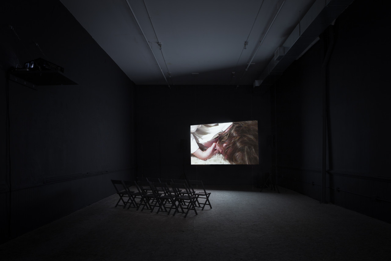 An empty dim-lit room with chairs with a projection on the wall. The projection features a close-up image of a person with wavy brown hair covering their face, lying facedown on a fluffy white carpet.