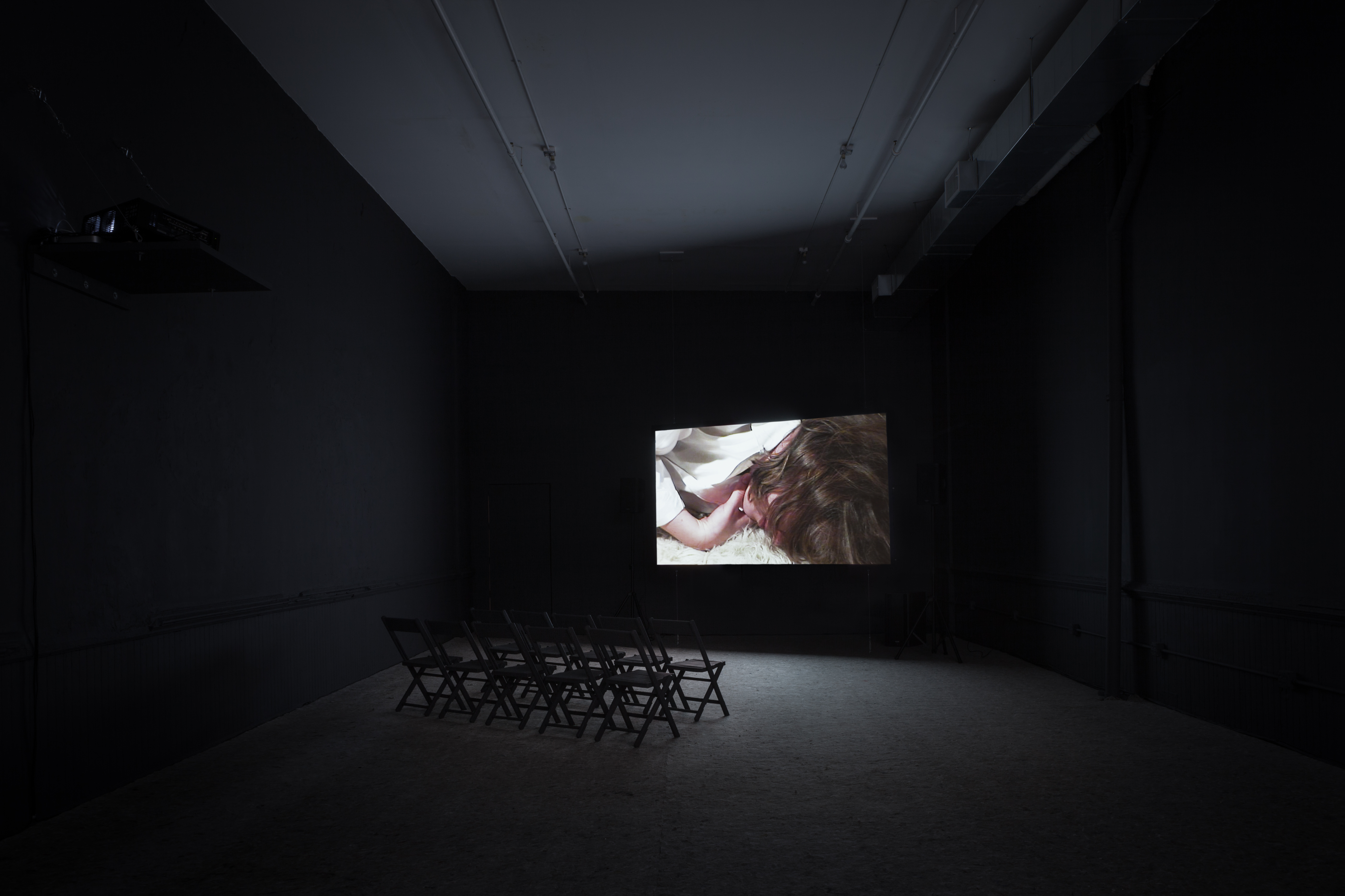 An empty dim-lit room with chairs with a projection on the wall. The projection features a close-up image of a person with wavy brown hair covering their face, lying facedown on a fluffy white carpet.