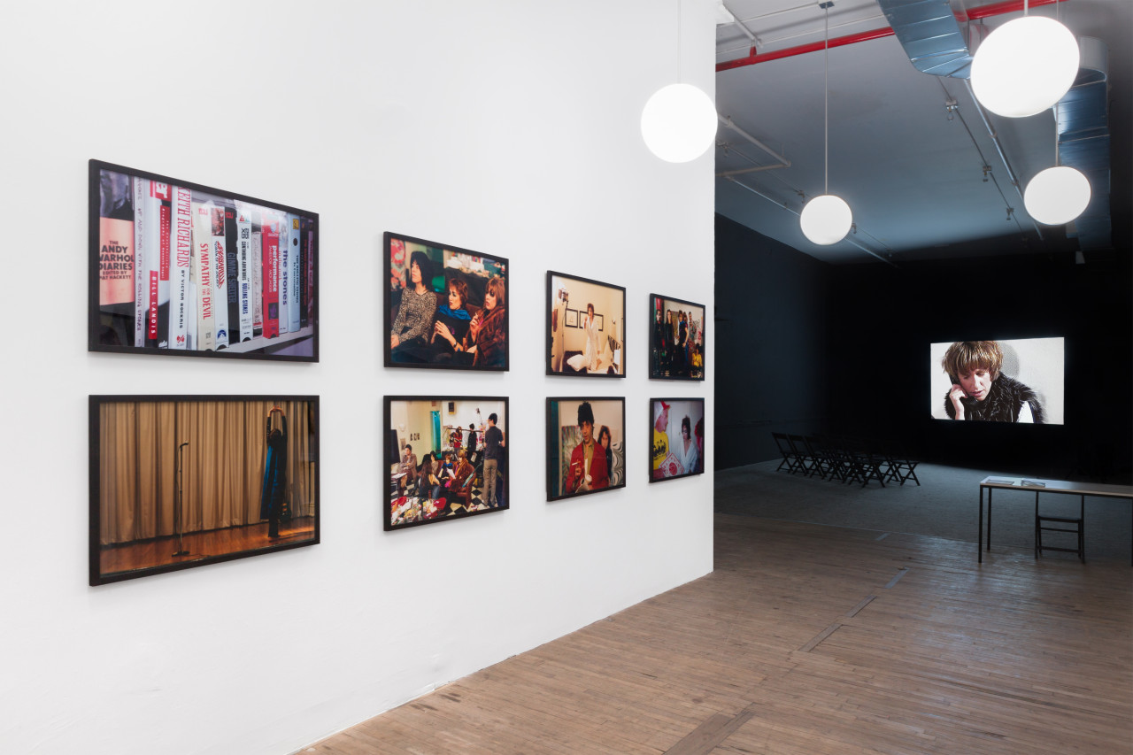 Eight large film stills are mounted on a wall, in two rows of four, leading into a dark hallway where a film projection is playing.