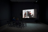 A photo taken on a diagonal of two people sitting on black chairs in a dimly lit room with a projection on the wall. The projection features a person with red hair looking at themself in a vanity mirror.