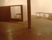 A color installation view of a gallery space with a white column, and three white tables.