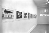 A row of images hangs on a white wall. The images are composed of simple shapes which are arranged to form scenes. The foremost image shows a house on a lawn.