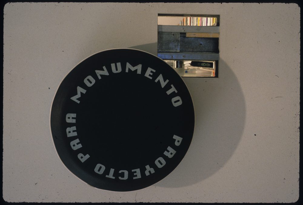 A large black disk installed on a white gallery wall. Bold gray text circling the circumfrence of the disk reads, "PROYECTO PARA MONUMENTO." Directly to the left of the disk, a square hole has been cut into the gallery wall. A horizontal metal bar and several planks of wood are visible through the hole.