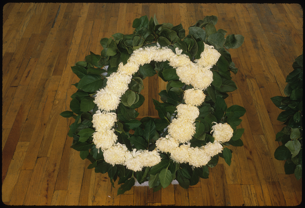 A flower arrangement consisting of white flowers and green leaves displayed on a slated, wooden floor. The arrangement is shaped such that it forms a lowercase, italicized letter "a."