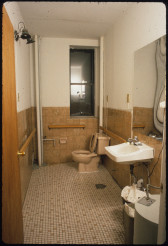 A wooden door opens on a view of a tiled, single-user bathroom. To the right of the room is a small, porcelain sink and warped mirror. Behind the sink is a tan toilet below a thin window.