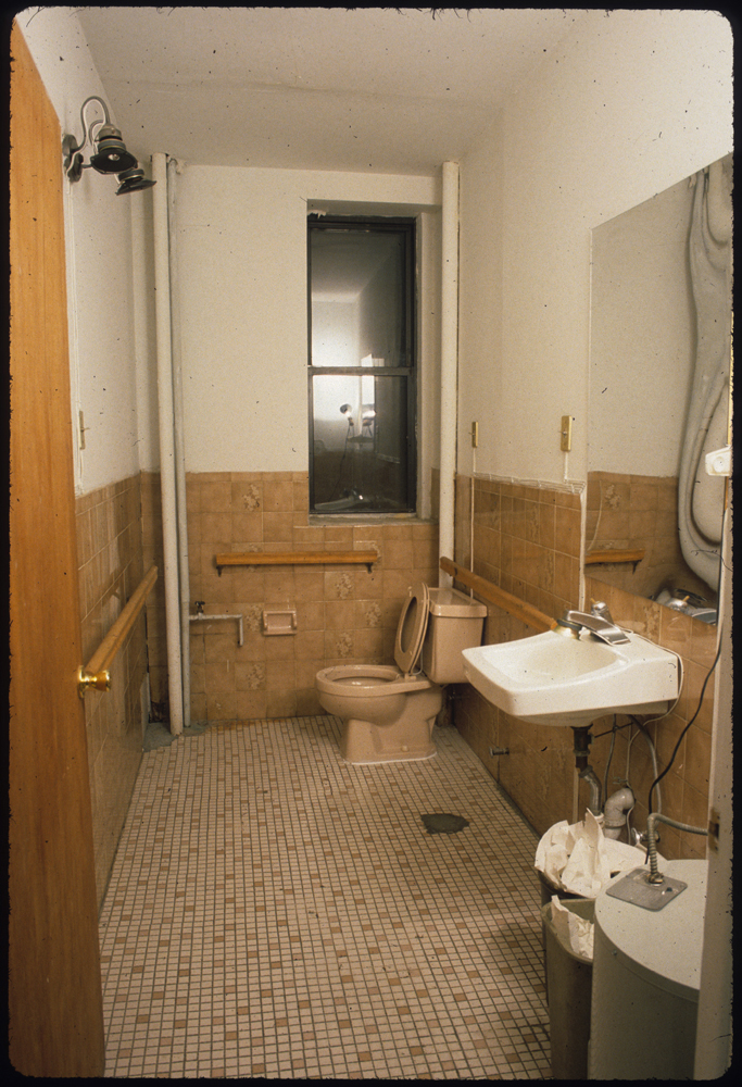 A wooden door opens on a view of a tiled, single-user bathroom. To the right of the room is a small, porcelain sink and warped mirror. Behind the sink is a tan toilet below a thin window.