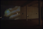 An image of a hand holding a yellow golf ball projected onto the wall of a dimly-lit gallery space. Several square holes have been removed from several spots on the gallery wall, through which a space on the other side of the wall is visible.