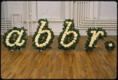 Five flower arrangements sit side-by-side on a gallery floor. The flowers have been arranged such that they form the shape of four italicized, lowercase letters and a period. From left to right, the arrangements spell 