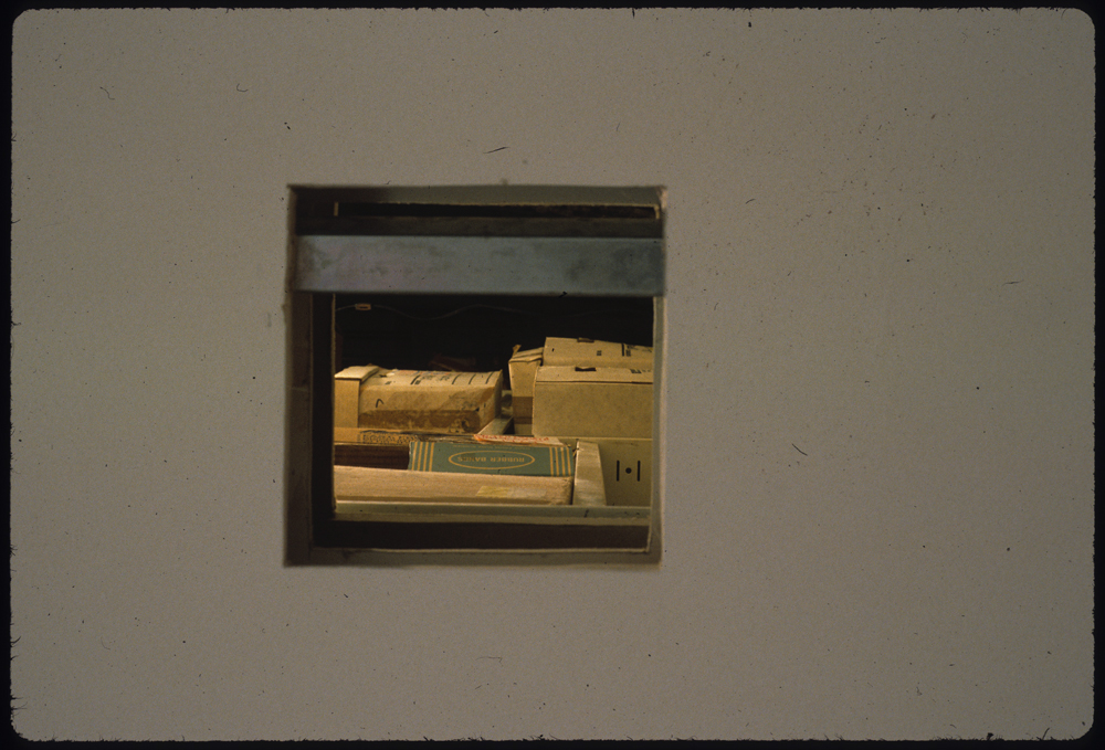 A square hole has been cut out of a blank, white gallery wall. Through the hole, several brown paper and cardboard boxes are visible.