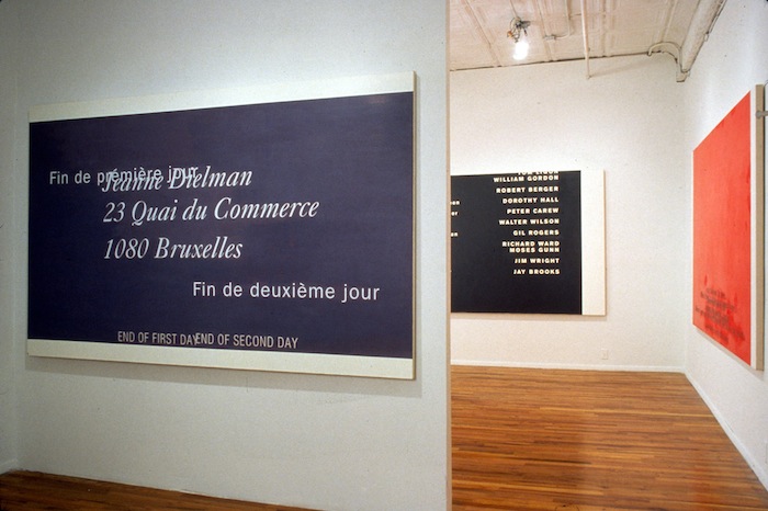 Three large, monotone paintings, each hung on its own white wall. A protruding white wall with a blue canvas and white text in French partially obscures a black canvas with white text. Perpendicular to that is a red canvas.