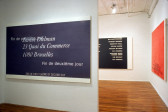 Three large, monotone paintings, each hung on its own white wall. A protruding white wall with a blue canvas and white text in French partially obscures a black canvas with white text. Perpendicular to that is a red canvas.