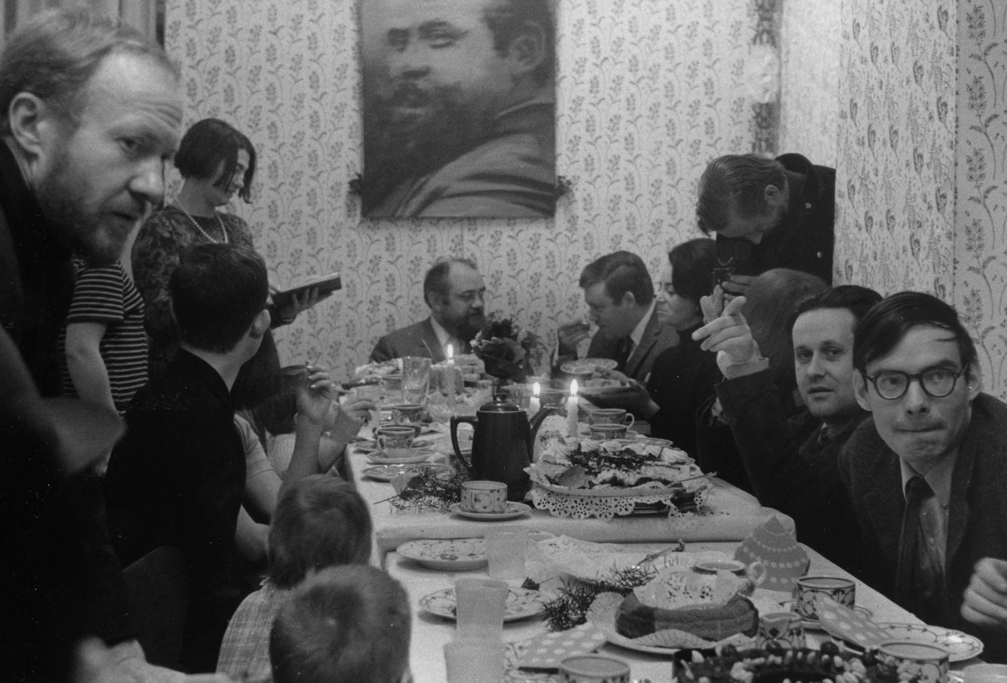 A black and white image of a crowd of people gathered around a dinner table. The walls of the room are patterned, and there is a portrait of a bearded man hanging on the wall above the guest at the head of the table.