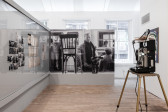 A wall-sized, black and white photograph of chair and man in front of a train car is adjacent to an angled wall displaying photographs and documents behind glass. In the right hand foreground is a 35mm projector.