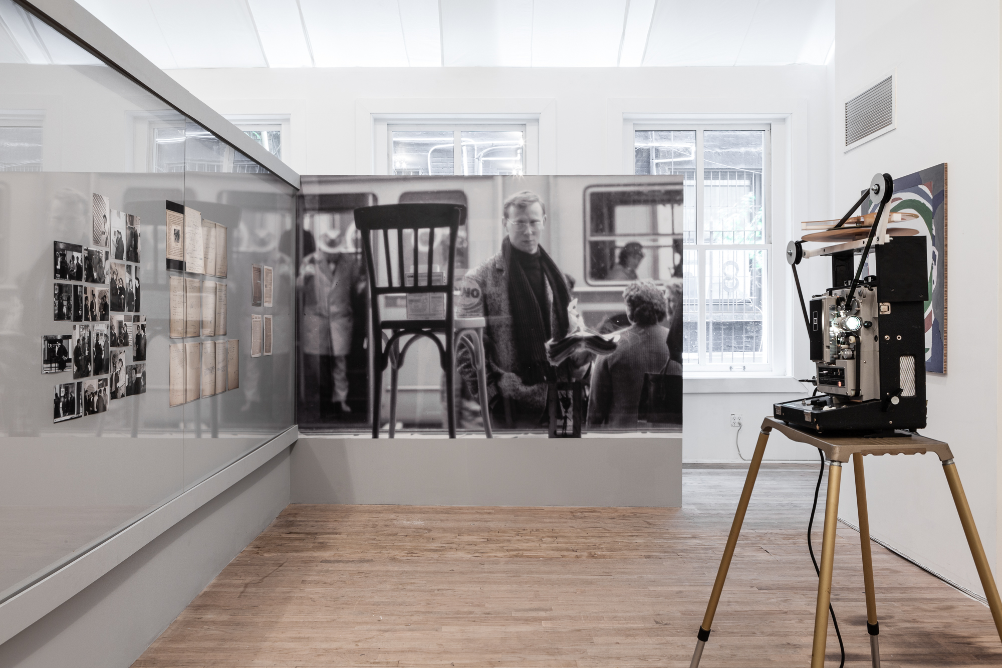 A wall-sized, black and white photograph of chair and man in front of a train car is adjacent to an angled wall displaying photographs and documents behind glass. In the right hand foreground is a 35mm projector.