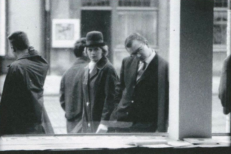A black and white image of a man looking at documents through a window on a street sidewalk. Behind him, a women in a hat looks at him. People are present walking on the street surrounding them.